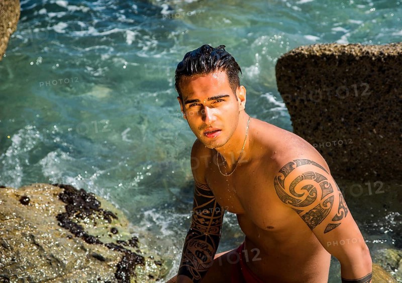 Attractive young shirtless athletic man standing in water by sea or ocean shore, wearing shorts, looking down to a side