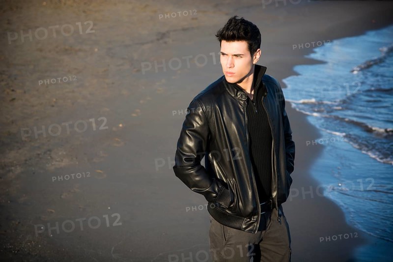 Handsome fashionable young man at the seaside on the beach with his black leather jacket looking away, large copyspace