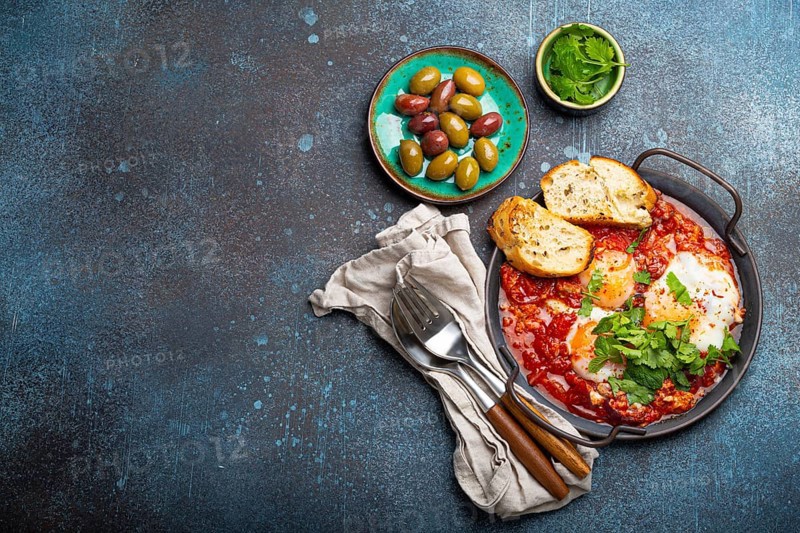 Middle Eastern and Maghrebi healthy dish Shakshouka made of eggs and tomato sauce served in pan with toasts, fresh cilantro and olives on rustic concrete background table from above, space for text, food photography