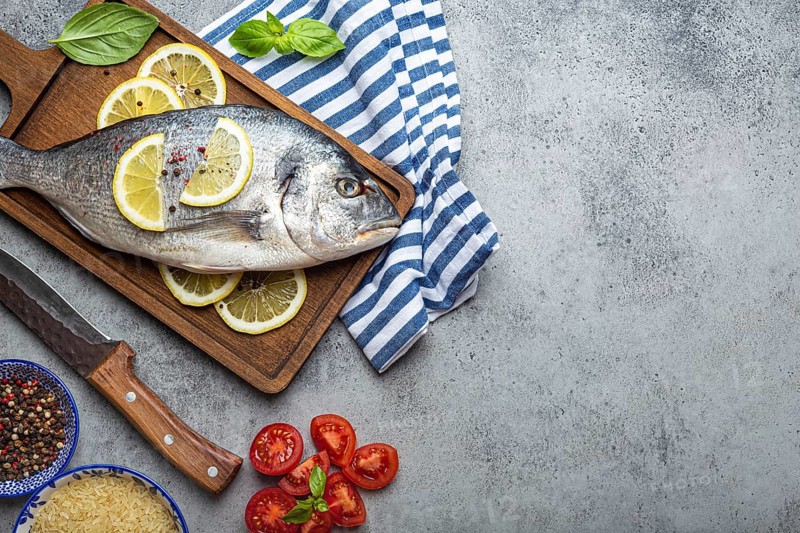 Raw fish dorado with ingredients lemon, fresh basil, cut cherry tomatoes, uncooked rice on wooden cutting board with knife on rustic stone background top view, cooking healthy fish dorado. Copy space, food photography, food photography