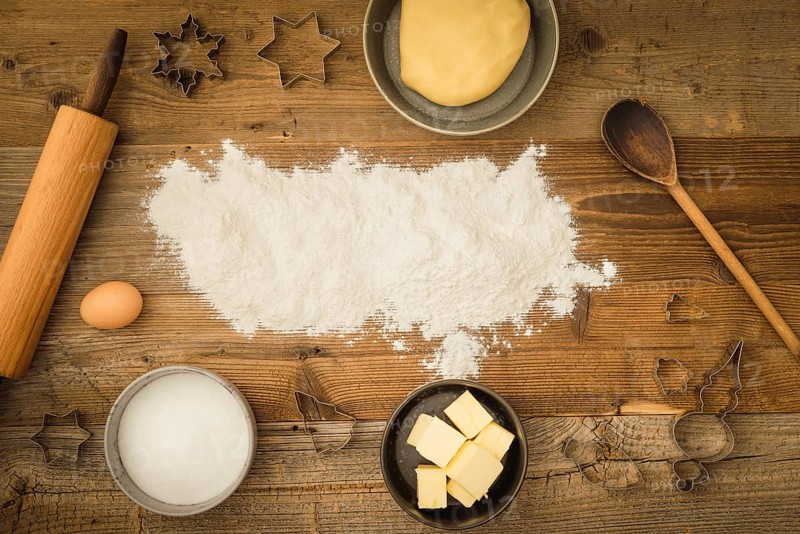 Collection of ingredients and utensils for baking Christmas biscuits with flour as text space in the centre of the picture on a dark wooden background