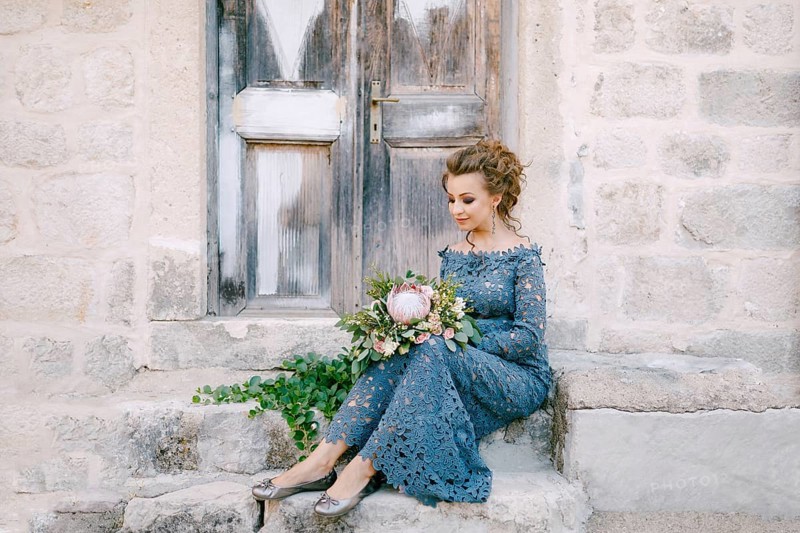A bride in a stylish blue dress with a bouquet in her hands sits on the steps near an old wooden door .