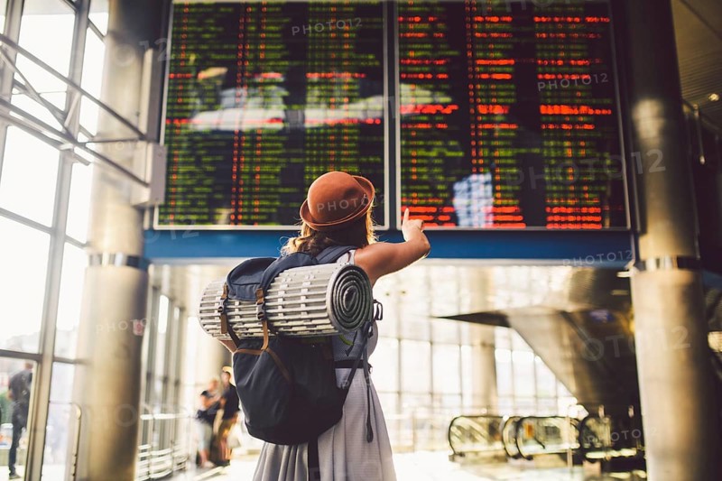 Theme travel public transport. young woman standing back in dress and hat behind backpack camping equipment for sleeping, insulating mat looks schedule on scoreboard airport station hand to the front
