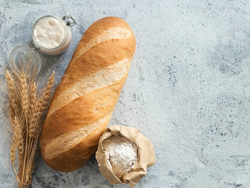 British White Bloomer or European sourdough Baton loaf bread on gray cement background. Fresh loaf bread and glass jar with sourdough starter, flour in paper bag and ears. Top view. Copy space