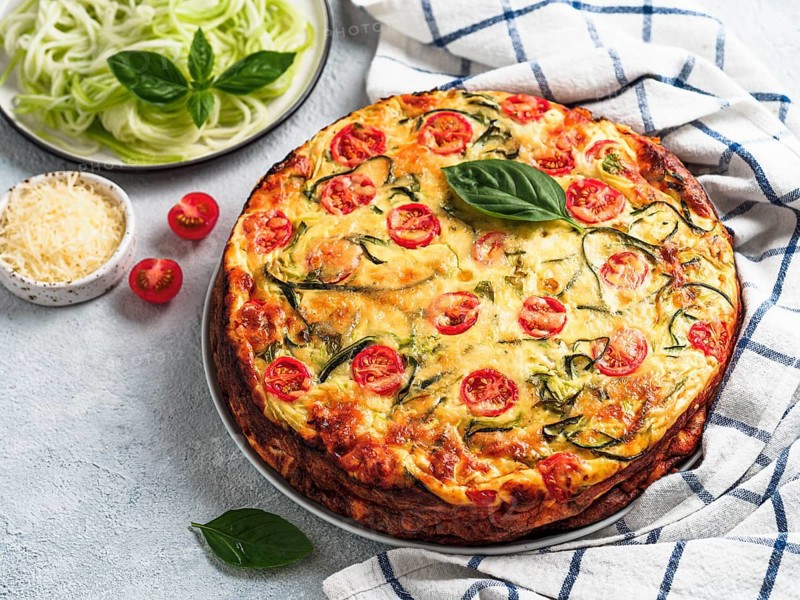 Zucchini pie and ingredients on gray background, copy space left. Delicious savory pie with zucchini, tomatoes, herbs and cheese. Idea and recipe for healthy baking and lot to harvest of zucchini