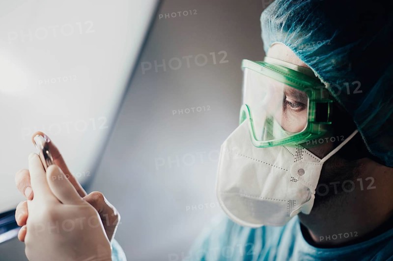 A doctor in green goggles examines a vial while wearing a mask
