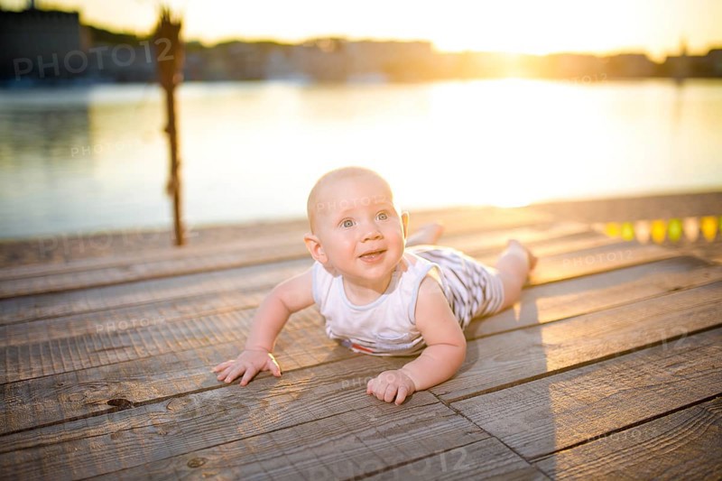 A child a boy, one year old, blond man lies on his stomach on wooden dock, pier in striped clothes, compound near pond on sandy beach against background of river in the summer at sunset of the day