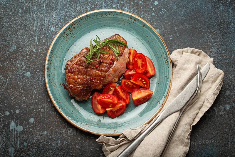 Delicious roasted duck breast fillet with golden crispy skin, with pepper and rosemary, top view on ceramic blue plate served with cherry tomatoes salad, rustic concrete rustic background, food photography, food photography