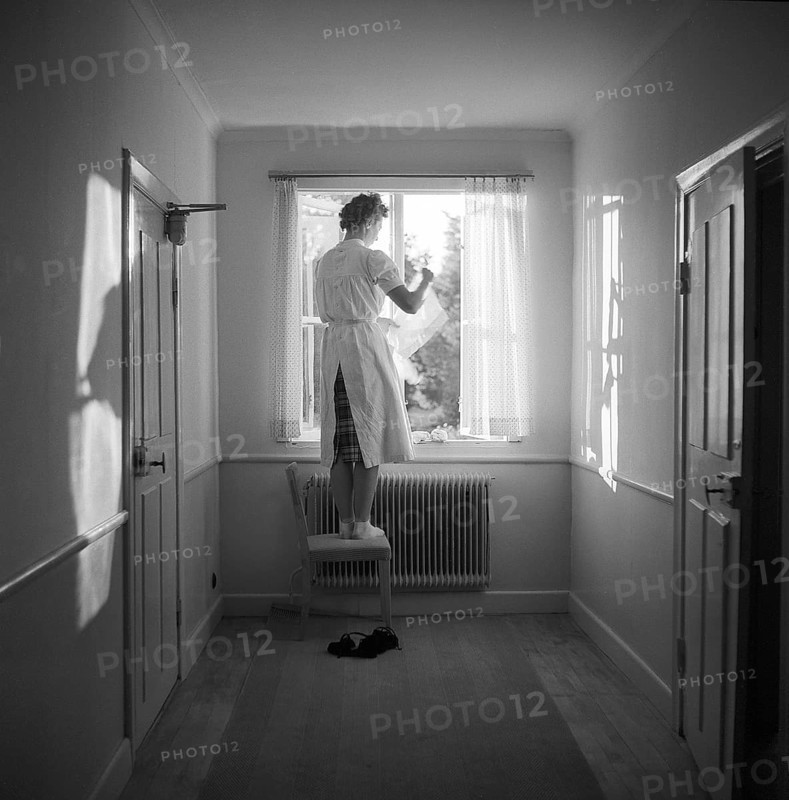 In the 1950s. A woman is seen at a window in a corridor standing barefoot on a chair to hang the window curtains up. 1955. Svahn