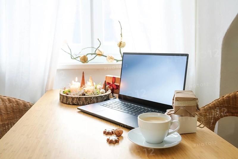 Laptop on a wooden desk by the window in an attic apartment. Gifts, advent candles, Christmas decoration and a coffee cup. Holiday online shopping, copy space, selected focus, narrow depth of field
