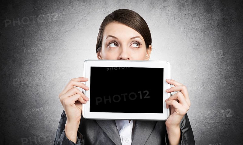 Businesswoman holding tablet computer with blank screen. Beautiful woman in business suit show tablet PC near her face. Corporate businessperson on grey wall background. Digital technology layout