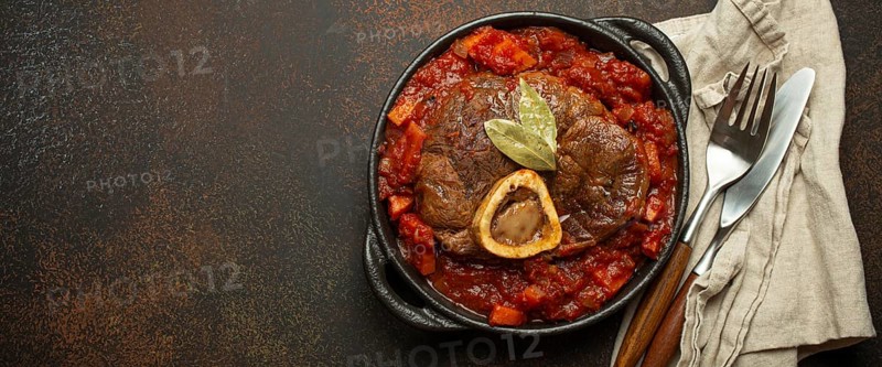 Traditional Italian dish Ossobuco all Milanese made with cut veal shank meat with vegetable tomato sauce served in black casserole pan top view on rustic brown background, copy space, food photography, food photography