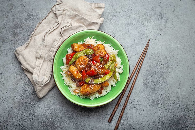 Asian sweet and sour sticky chicken with vegetables stir-fry and rice in ceramic bowl with chopsticks top view on gray rustic stone background, traditional Asian dish, food photography, food photography