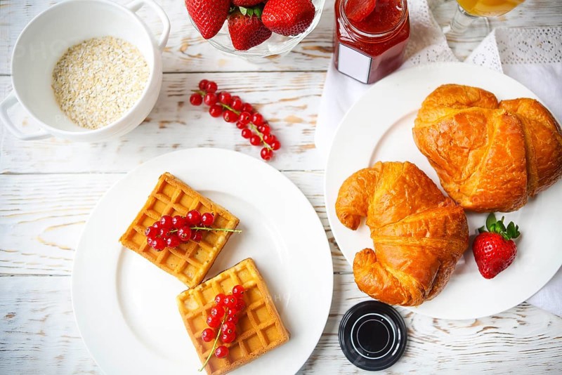 Waffles with red currant jam and berries on a white plate, croissants, orange juice and oat flakes oatmeal on the wooden background