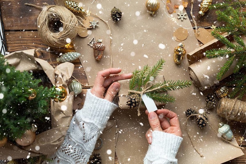 Hands pack a gift for Christmas and new year in eco-friendly materials: kraft paper, live fir branches, cones, twine, tied with a bow. Tags with mock up, natural decor, hand made, flatly. Festive mood
