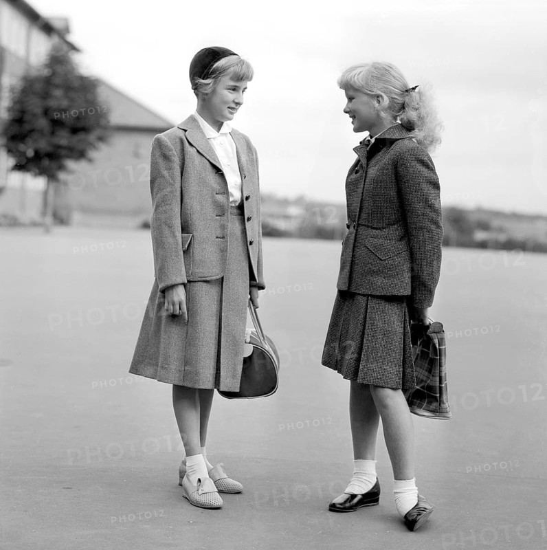 Girls in the 1950s. Two teenage girls dressed in outfits typical of the decade. Typical schoolgirls holding their schoolbags standing outside the school building on the schoolyard. 1954. Conard ref 2722