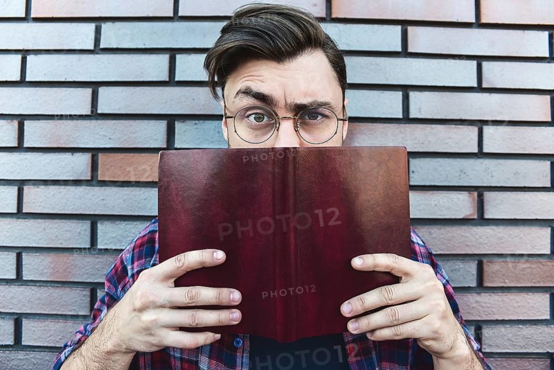 Funny face. Young hipster guy wearing eyeglasses with picked up one browe looking at camera and holding a book or note book on brick wall background