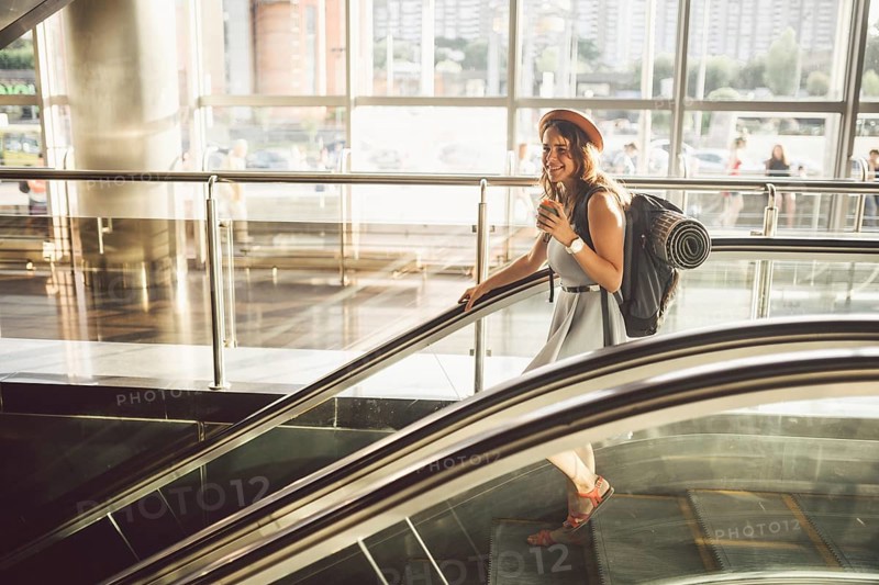 Portrait Smiling Woman Wearing Hat In Airport At Escalator. people travelling with hand luggage. Theme tourism and transport. Caucasian girl with coffee in airport terminal on an escalator