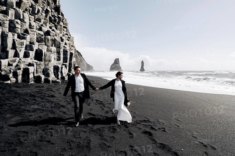 Destination Iceland wedding. A wedding couple walks along the sandy black beach of Vik, near the basalt rock, in the form of pillars. The bride and groom hold hands and walk along the shore