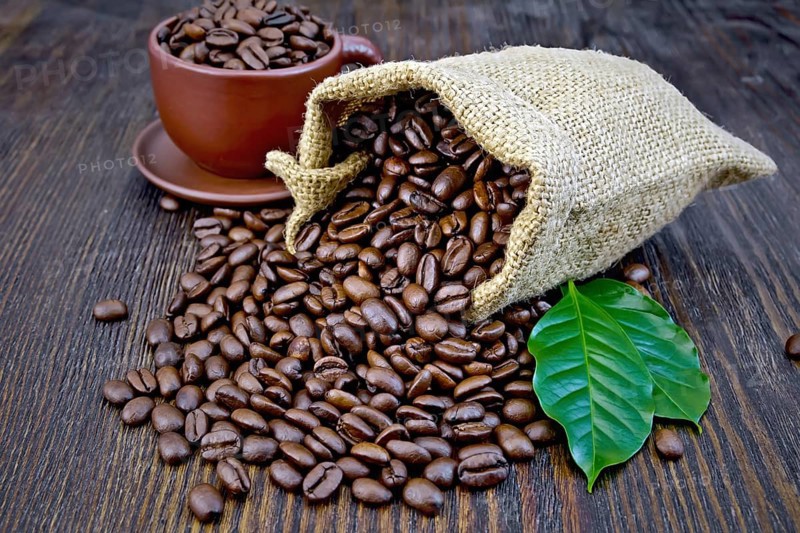 Bag of black coffee beans, brown cup and green leaves on a wooden board background
