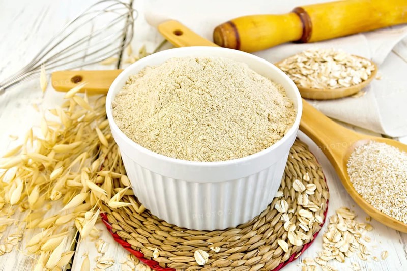 Oat flour in white bowl, oatmeal and bran in wooden spoon, stalks of oats on the background of wooden boards