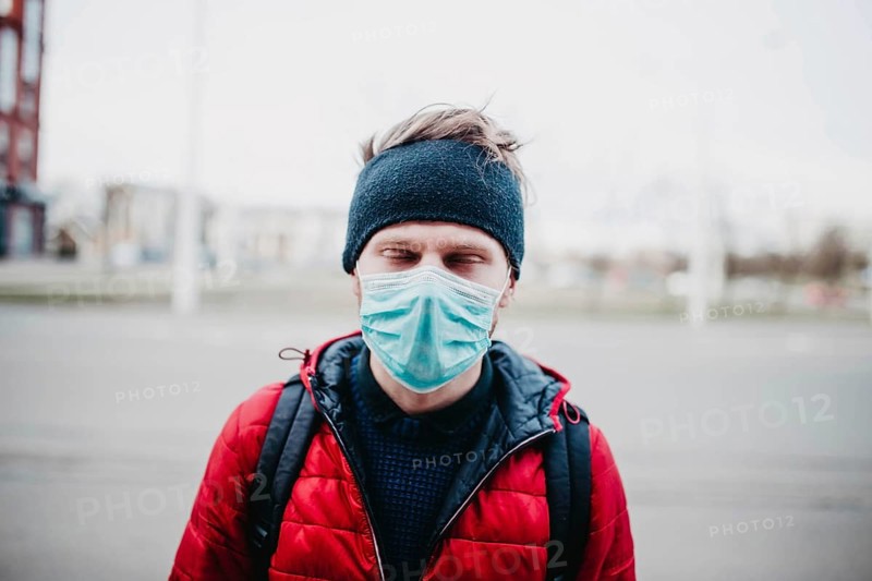 Man in a red jacket and face mask outdoors, with city buildings in the background