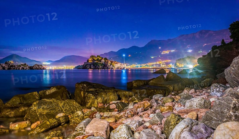 Stones on the coast of Adriatic sea near illuminated Sveti Stefan at sunset, Montenegro, Europe
