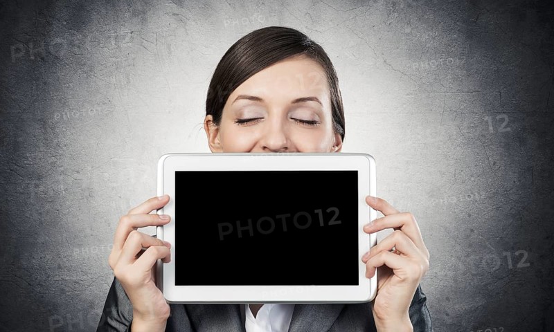 Businesswoman holding tablet computer with blank screen. Beautiful woman with closed eyes showing tablet PC near her face. Corporate businessperson on grey wall background. Digital technology
