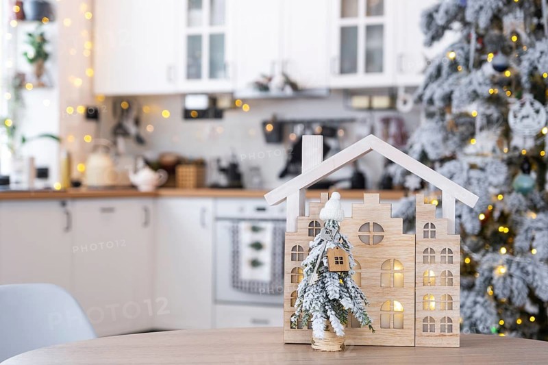 Key on Christmas tree and tiny house on cozy home with Christmas decor on table of festive white kitchen. Gift for New Year. Insurance, moving to new house, mortgage, rent and purchase real estate