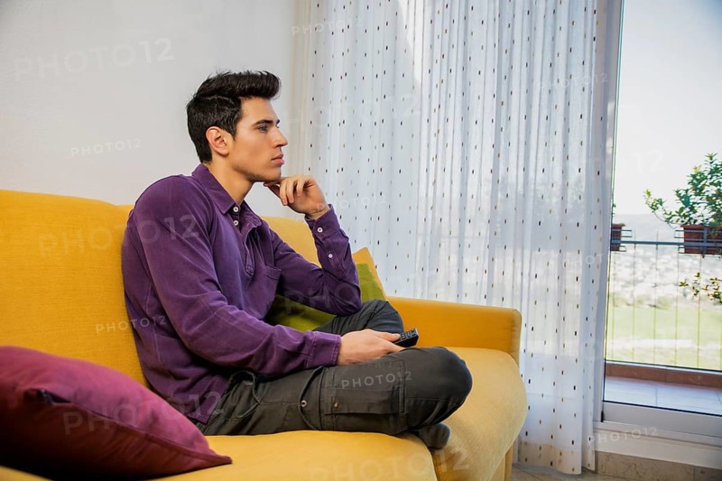 Young man sitting on couch watching television changing the channel with the remote control