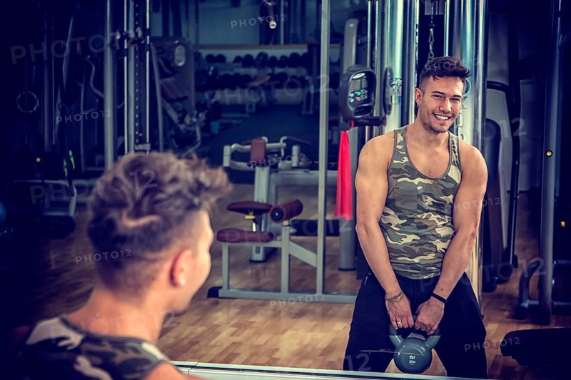 Attractive young athletic man exercising in gym, working out using kettlebell, looking at camera with a smile