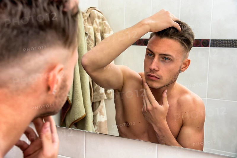 Handsome man looking at his face in mirror, checking hair and hairline