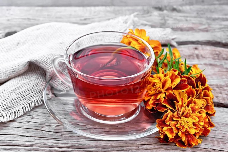 Marigold herbal tea in a glass cup and saucer, fresh flowers, sackcloth napkin on wooden board background