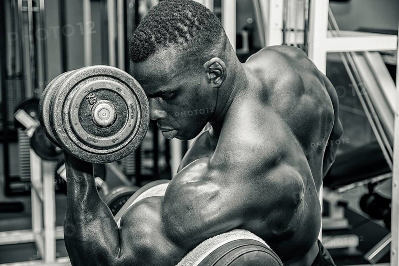 Hunky muscular black bodybuilder working out in gym, exercising biceps with dumbbell. Black and white photo