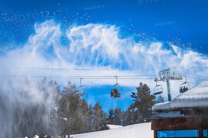 Snow cannon spreading fresh snow powder on a ski slope. Skiers riding up on chair lift. Winter ski holidays in El Tarter, Andorra, Pyrenees Mountains, Europe