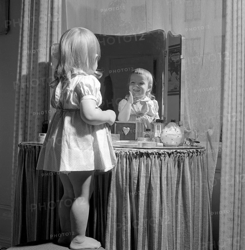 In the 1950s. A little girl is standing in front of her mothers makeup table and mirror and puts on some kind of beauty cream on her face, looking happy. Sweden 1951 Conard ref 1708