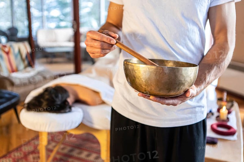 Massage therapist standing with a singing bowl in hands and giving a sound massage, to his client lying on the massage bed at the background