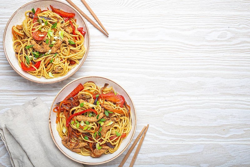 Two bowls with Chow Mein or Lo Mein, traditional Chinese stir fry noodles with meat and vegetables, served with chopsticks top view on rustic white wooden background table, space for text, food photography, food photography