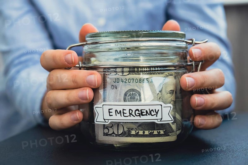 Female hands Saving Money In Glass Jar filled with Dollars banknotes. EMERGENCY transcription in front of jar. Managing personal finances extra income for future insecurity background
