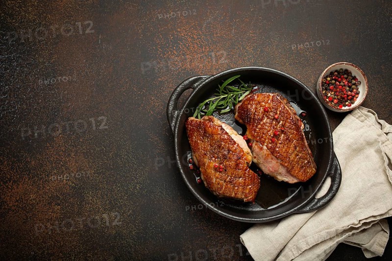 Two roasted duck breast fillets with crispy skin, with pepper and rosemary, top view in black cast iron pan with knife, dark brown concrete rustic background, space for text, food photography, food photography