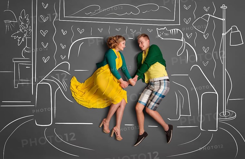 Happy valentines love story concept of a romantic couple sitting on a sofa and holding hands against chalk drawings background of a living room