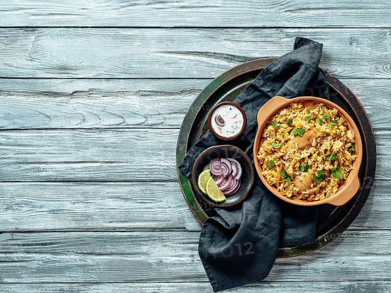 Pakistani food - biryani rice with chicken and raita yoghurt dip. Delicious hyberabadi chicken biryani over tray on gray wooden background. Top view or flat lay. Copy space for text or design