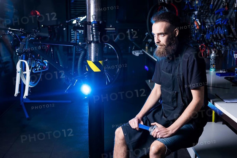 Portrait of small business owner of young man with beard. Guy bicycle mechanic workshop worker sitting with tool in his hand in a working black clothes in an apron in the background of a bicycle shop