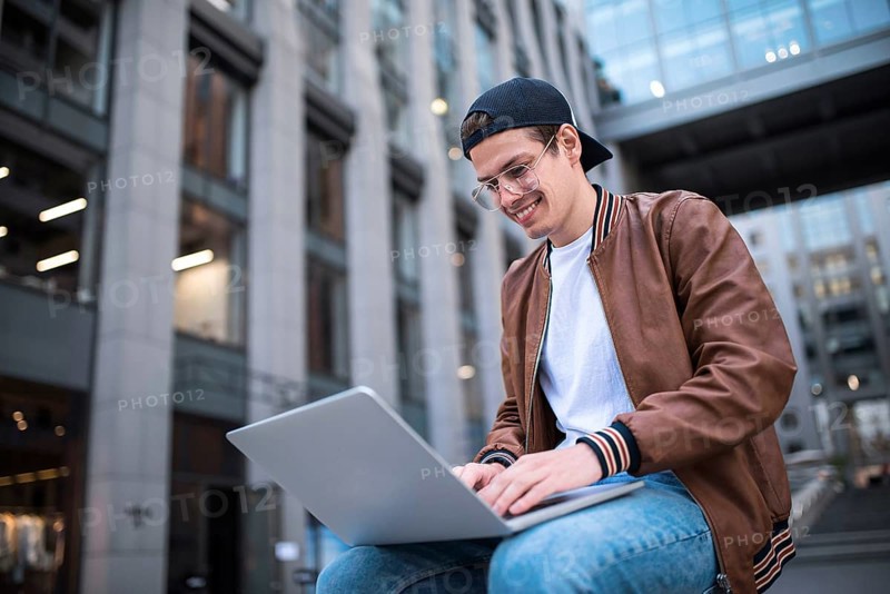 Happy handsome student looking at computer screen while typing something on laptop keyboard during work break outdoors, teenager looking at the notebook sitting at street