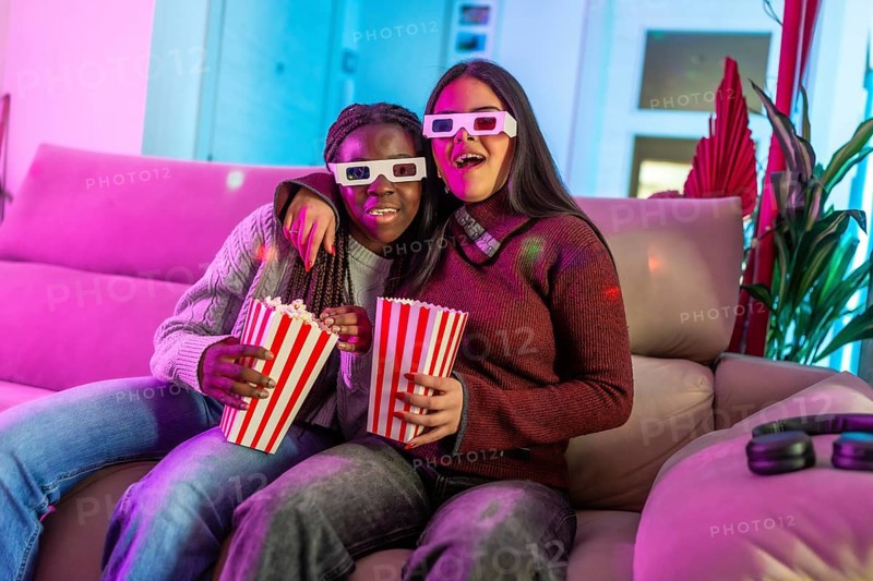 Two girls wearing 3d glasses enjoying a movie night at home, eating popcorn from striped boxes and having fun on the sofa