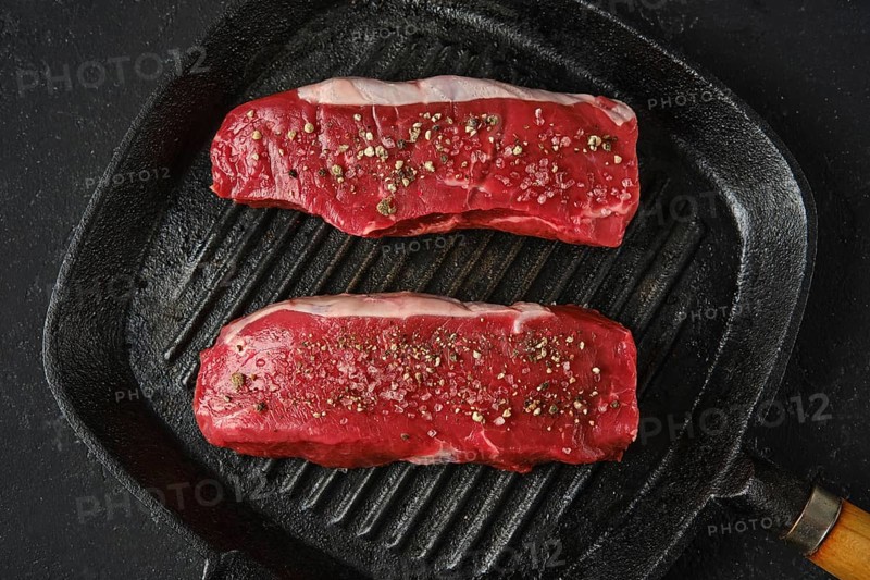 Top view of two thick cuts of raw beef steaks on a cast iron skillet. The marbled texture of the meat is seasoned with sea salt and pepper