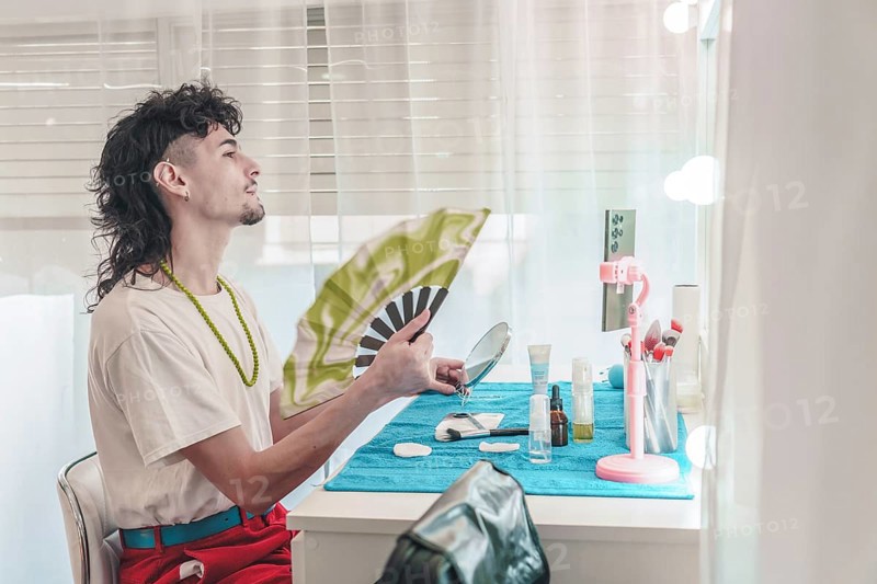 Young influencer with mullet hairstyle is cooling himself with a hand fan while applying makeup in front of a mirror in his bedroom, showing diversity and inclusion