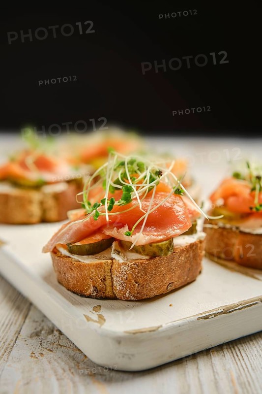 Open-faced sandwiches are arranged on a wooden platter, featuring layers of smoked salmon, pickles, and sprouts. This appetizing display highlights vibrant colors and fresh ingredients.