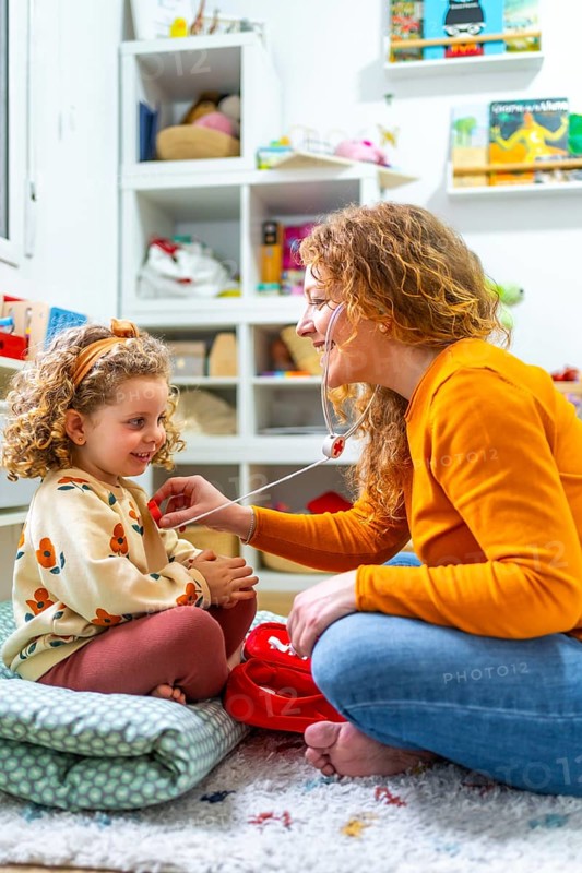 Vertical photo of a caucasian mother listening the beat of daughter heart with fake stethoscope playing nurses and doctors at home