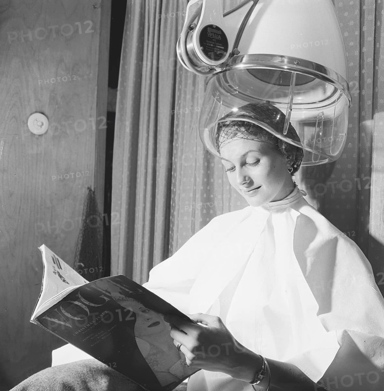 Hair dryer in the 1950s. A woman is sitting comfortably and reading Vogue magazine, while drying her hair under a drying hood. Sweden March 1954